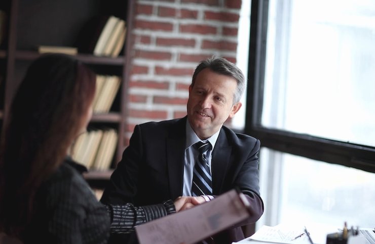 A man in a suit handing a folder to a woman across a desk with a laptop and bookshelf behind them
