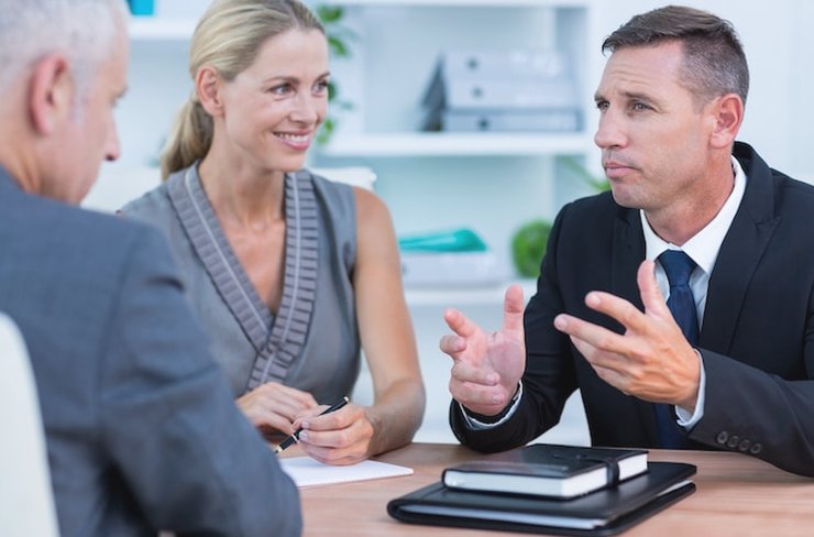 Two men and a woman in business clothes talking across a desk while she takes notes