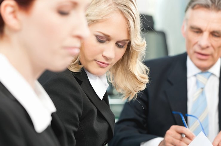 Three colleagues in business clothes, two reading papers and an older man signing a page at a meeting table