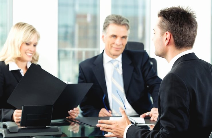 Four people in dark suits at a conference table in a bright office, one of them speaking and gesturing