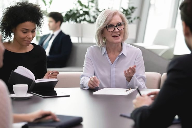 Colleagues talking around a table with notebooks and coffee, a grey-haired woman with glasses speaking
