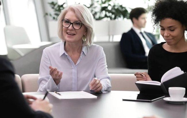 A grey-haired woman with glasses speaking to colleagues around a table with notebooks and a cup of coffee