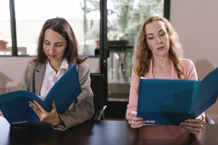 Two women at a dark table, each reading an open blue folder