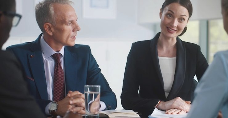 A woman and a man in suits seated at a table, smiling and talking, with a glass of water in front of them